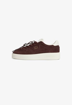 Baskets basses en daim marron foncé avec semelle blanche, détail blanc au talon, lacets à motifs et logo discret sur le côté et la semelle.