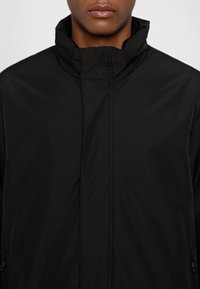 Giacca impermeabile nera con colletto alto e chiusura con zip. Texture liscia, design minimalista e tasche laterali.