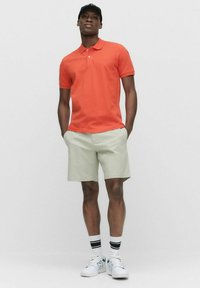 Orange polo shirt, lys grønne shorts, hvide sneakers med teal-accenter og sort-hvide stribede sokker, stående mod en neutral baggrund.