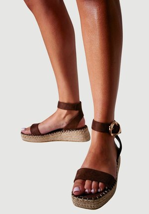 Beine in braunen Wildleder-Plattform-Sandalen mit Knöchelriemen und großen goldenen Schnallen vor neutralem Hintergrund.