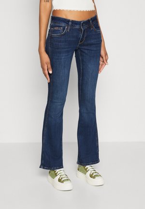 Mörkblå utsvängda jeans i denim, med framfickor, dragkedja och kontrasterande sömmar. Bärs med gröna sneakers.