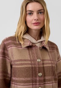 Brązowo-różowy wełniany płaszcz w kratę z kołnierzem, zapięciem na guziki oraz beżowym, ocieplanym kapturem. Charakteryzuje się dużymi guzikami i miękką teksturą.