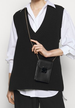 Zwart gebreid vest met een V-hals ontwerp gedragen over een wit shirt, gecombineerd met een zwarte leren crossbody tas met een kettingriem en sluiting.
