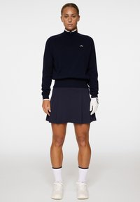 Sininen vetoketjullinen villatakki, jossa on valkoinen logo, yhdistettynä siniseen laskostettuun hameeseen. Valkoiset hanskat ja kengät, erottuvat valkoiset urheilusukat.