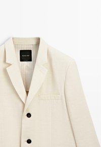 Beżowa marynarka jednorzędowa z wciętymi klapami, dwoma czarnymi guzikami i kieszenią na piersi, oznaczona marką Massimo Dutti.