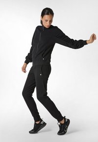 Tuta sportiva nera realizzata in tessuto morbido, caratterizzata da una giacca con zip e polsini a costine e pantaloni tapered abbinati con tasche laterali.