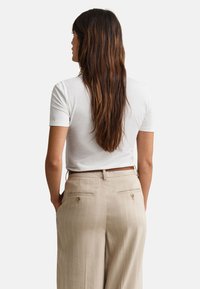 Žena s dugom smeđom kosom stoji, nosi bijelu košulju kratkih rukava uvučenu u bež hlače s tigrastim uzorkom i smeđi pojas.