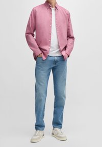 Hombre de pie con las manos en los bolsillos, llevando una camisa de botones rosa, una camiseta blanca, jeans azules y zapatillas de color claro frente a un fondo liso.