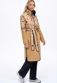 Prošívaný trenčkot v béžové barvě s černými puntíky, dlouhými rukávy, páskem v pase a kontrastním černým lemováním; vyrobený z prošívané látky.