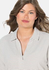 Camicia colletto bianca con strisce orizzontali marroni, caratterizzata da una chiusura a bottoni e una vestibilità comoda. Il tessuto sembra morbido e leggero.