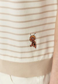 Haut en tricot rayé avec des couleurs beige doux et crème. Présente un personnage de dessin animé brodé et amusant à l'ourlet. Ceinture côtelée texturée.