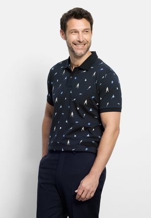 Mann lächelt, trägt schwarzes Polohemd mit blauen und weißen Tennis-Symbolen und dunkle Hose, steht vor einem schlichten hellen Hintergrund.