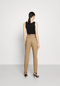 Frau mit dunklen Haaren, die nach hinten gebunden sind, trägt ein schwarzes ärmelloses Top, beige Hochbundhosen und schwarze High-Heels, und steht mit dem Rücken zu uns.