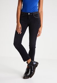 Jeans neri a taglio stretto, con borchie decorative sui bordi delle tasche, abbinati a stivali neri lucidi alla caviglia e a una maglia blu.