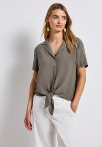 Camicetta a maniche corte di colore verde oliva in tessuto leggero, con colletto e orlo anteriore legato. Abbinata a pantaloni bianchi, modello che indossa orecchini.