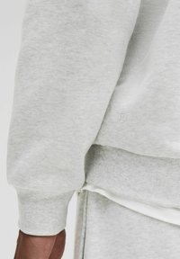 Grauer Pullover mit gerippten Bündchen und Saum. Weicher, strukturierter Stoff mit kleinem Logo am Saum. Geschichteter Stil mit weißem, sichtbarem Unterteil.