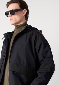 Sort hættejakke med lynlås, tekstureret stof og kontrasterende paneler. Bæres over en grøn turtleneck. Sorte rektangulære solbriller.