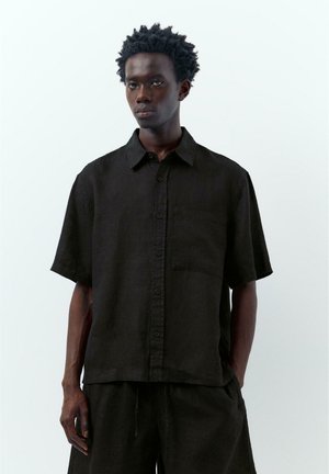 Jeune homme aux cheveux courts et bouclés portant une chemise noire ample à manches courtes boutonnée et un pantalon noir assorti sur un fond uni.