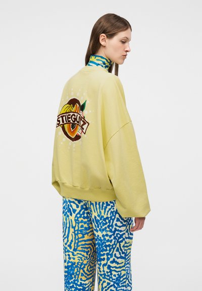 Femme portant un sweat-shirt oversize jaune pâle avec un motif de fruits colorés et le texte "STIEGLITZ" au dos, associé à un pantalon à motifs bleus et jaunes.