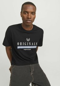 Camiseta de algodón negra con estampado "ORIGINALS" en blanco y líneas de acento azul. Manga corta y cuello redondo. Combinada con jeans oscuros.