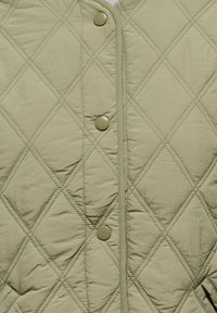 Gilet matelassé en tissu vert pâle présentant un motif en losange, des boutons-pression en plastique ronds et un aspect légèrement brillant. Texture lisse et coupe ajustée.