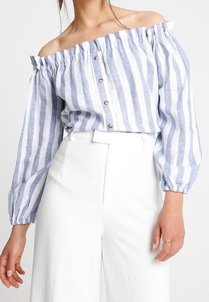 Femme portant une blouse à rayures verticales bleu et blanc avec épaules dénudées et boutons, associée à un pantalon blanc taille haute et des bagues aux doigts.