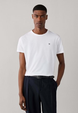 Man draagt een effen witte T-shirt met korte mouwen met klein logo op de borst, zwarte riem en donkere geplooide broek, staand tegen een lichte achtergrond.