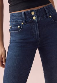 Jeans in denim blu scuro con vita alta, dotati di bottoni dorati, quattro tasche e una trama liscia. Visto da vicino.
