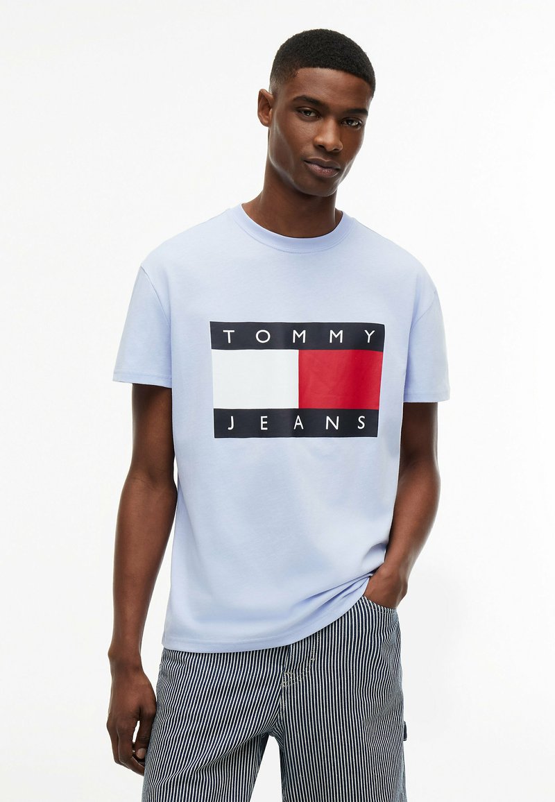 Jeune homme portant un t-shirt Tommy Jeans bleu clair avec un logo rouge et blanc, associé à un pantalon rayé noir et blanc, debout avec une main dans la poche.