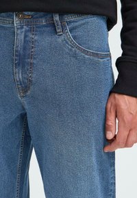 Jeans din denim de un albastru deschis, cu o croială clasică, având buzunare frontale, cusături contrastante și o închidere cu nasture la talie.