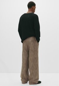 Mann, der einen schwarzen Strickpullover und weite, braune Cordhosen trägt, steht mit dem Rücken zu einem schlichten weißen Hintergrund.