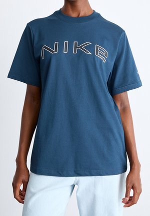 Persoon draagt een blauwe Nike T-shirt met een groot wit omlijnd logo over de borst en lichtblauwe spijkerbroek, staand tegen een witte achtergrond.