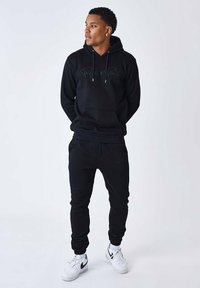 Zwarte hoodie met trekkoorden en logo, gecombineerd met zwarte joggingbroek. Witte sneakers maken de outfit compleet. Glad textuur overal.