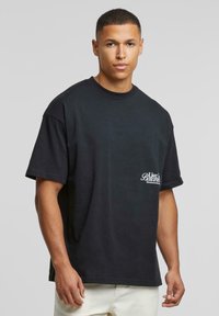 Černé oversized tričko vyrobené z bavlny s krátkými rukávy, s bílým logem na levém hrudníku a klasickým kulatým výstřihem.