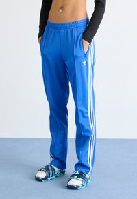 Oseba v svetlo modrih Adidas trenirkah z belimi črtami, ki stoji na modri podlagi, z rokama v žepih, obuta v modro-zelene copate s sokni.