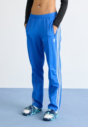 Oseba v svetlo modrih Adidas trenirkah z belimi črtami, ki stoji na modri podlagi, z rokama v žepih, obuta v modro-zelene copate s sokni.