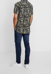 Kortärmad skjorta med ett mörkt mönster, prydd med geometriska former och detaljer. Bärs tillsammans med mörkblå jeans och vita sneakers.