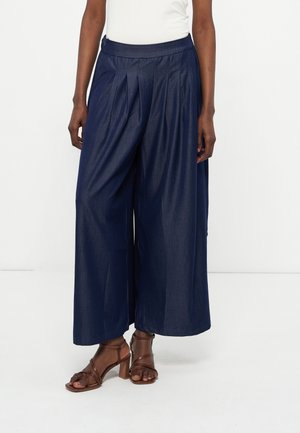 Persona che indossa pantaloni pieghettati larghi blu scuro e sandali con tacco marroni su uno sfondo bianco.