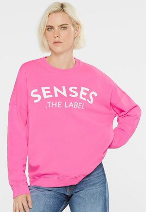 Bright pink sweatshirt with white text reading "SENSES .THE LABEL," featuring a crew neck, relaxed fit, and ribbed cuffs.