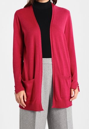 Magenta öppen cardigan med långa ärmar, två framfickor och ribbade detaljer. Bärs över en svart polotröja och grå byxor.