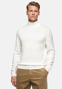 White turtleneck sweater made of a soft knit fabric, featuring long sleeves and ribbed hem. Worn with beige corduroy pants.