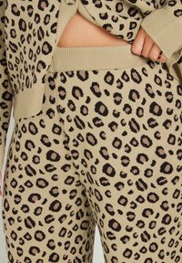 Beige stickad loungekläder som har ett mönster av leopardtryck i mörkbrunt och taupe. Tyget har en mjuk, texturerad yta.