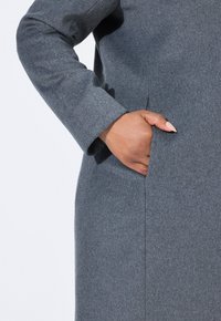 Cappotto di lana grigio con un design elegante, dotato di una tasca laterale e un taglio dritto. La texture sembra morbida e leggermente spazzolata.