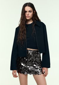 Schwarze, kurz geschnittene Jacke mit einem strukturierten Kragen über einem enganliegenden schwarzen Oberteil, kombiniert mit einem reflektierenden schwarzen Pailletten-Minirock mit gewellten Kanten.