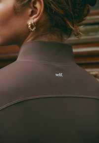 Pruun, sobiv ülaosa kõrge kaelusega ja peene tikandiga. Seljal on logo "self.". Vasakul on hästi nähtav suur kuldne rõngas kõrvarõngas.
