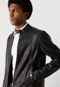 Schwarze Lederjacke mit Stehkragen, Reißverschluss, Seitentaschen und tonal abgesteppten Nähten. Verfügt über eine glatte Textur und ein minimalistisches Design.