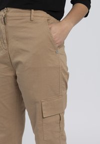 Pantaloni cargo beige realizzati in tessuto leggero, caratterizzati da numerose tasche, una vestibilità comoda e un logo visibile sulla tasca anteriore.