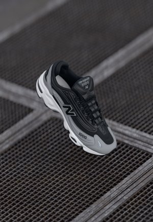 Baskets New Balance 1000 noires et grises suspendues au-dessus d'un sol en grille métallique avec des accents blancs et des lacets noirs.