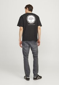 Svart bomulls t-skjorte med et sirkulært tre-design på baksiden, kombinert med grå denim jeans og svarte sneakers med hvite detaljer.
