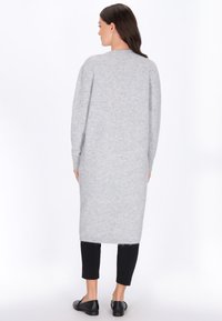 Femeie îmbrăcată cu o cardigan lung de culoare gri deschis, pantaloni negri scurți și loafers negri, stând cu spatele în fața unui fundal alb.